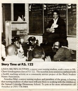 Black and white newspaper clipping of a student sitting in the front of a classroom reading a book to a group of students sitting on the floor.