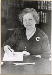 Photograph of a woman with curly hair sitting at a table with a pen and paper, she is smilin at the camera
