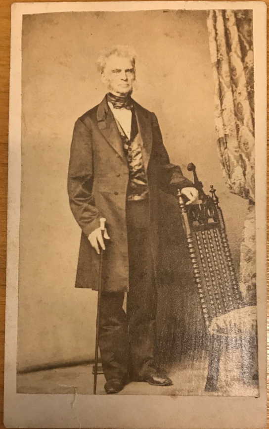 Sephia photograph of an older man leaning against a chair, he is wearing a long coat, bow tie, and vest.