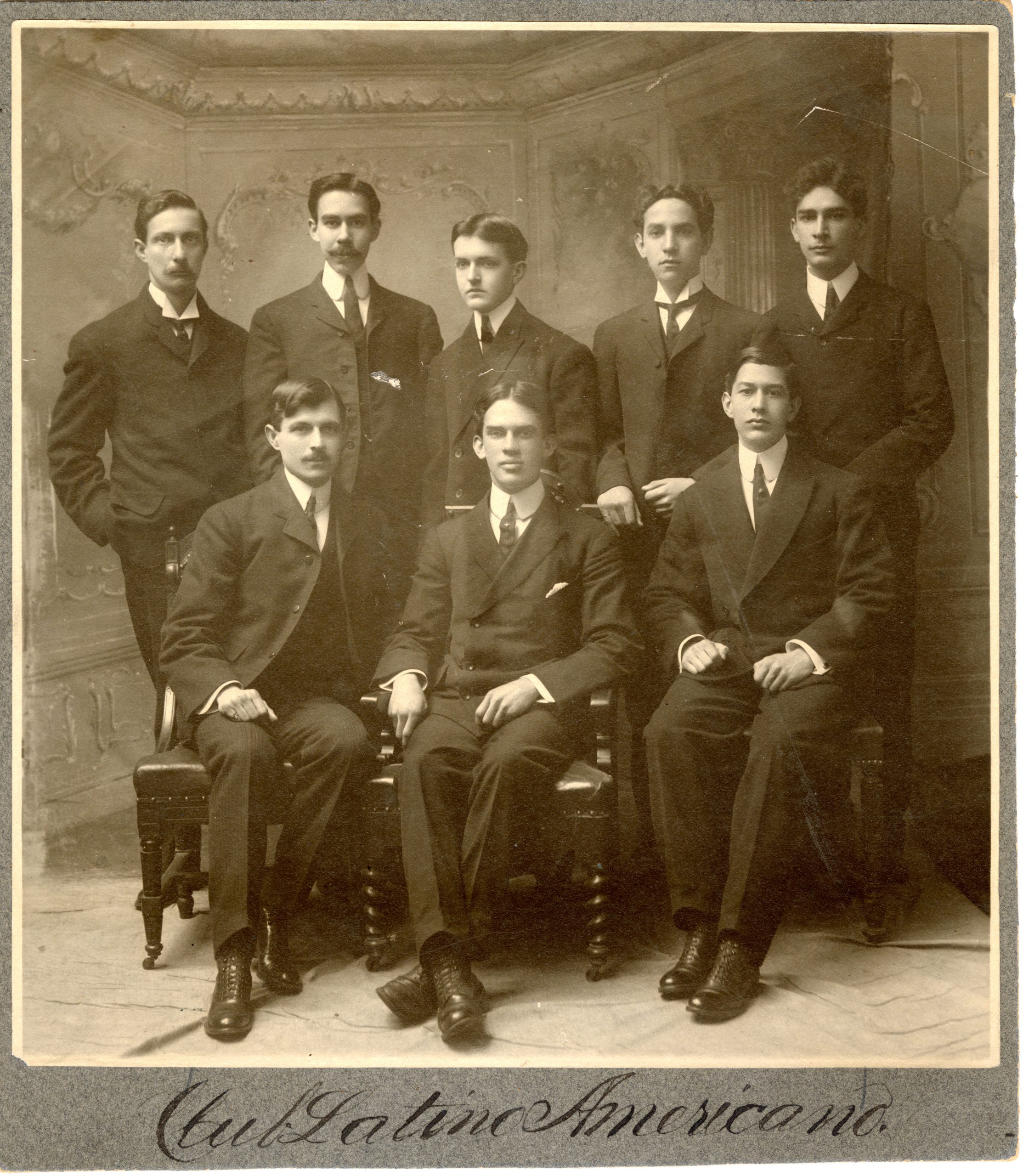 Sepia photograph of a group of 8 men, five standing behind three seated in chairs. Caption on the bottom handwritten says Club Latino Americano.