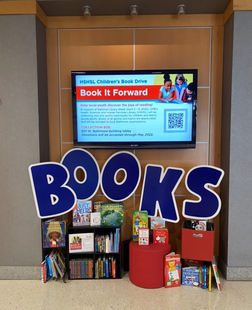Photograph with books on shelves and in boxes in the foreground, a large sign that says "books" and a screen advertising the book it forward drive in the back.