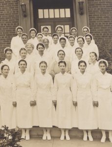 Black and white photograph of a group of posed women in nursing attire standing on stairs in front of a brick building.