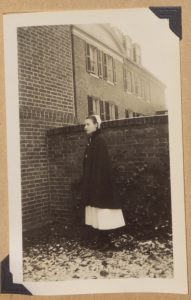 Black and White photograph of a women in coat and nurses dress and cap in front of a brick building.