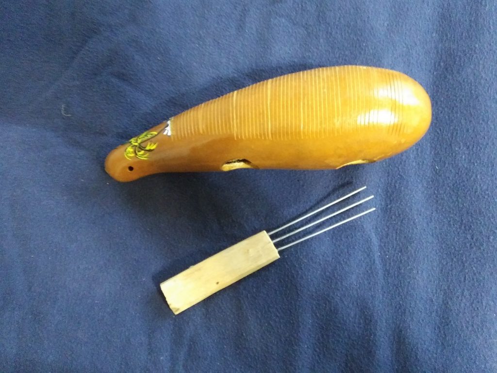 Photograph of a güiro and scraper, the güiro is a wooden gourdlike shaped insrument with various length scrapes across its top and two finger-sized holes on the side. The scraper has a handle and three metal prongs.