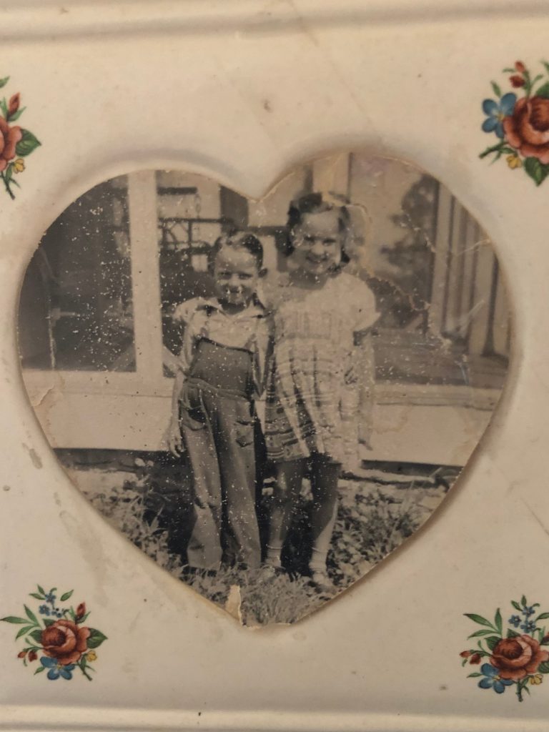 Sepia photograph of a little boy in overalls with a little girl in a dress with her arm around him