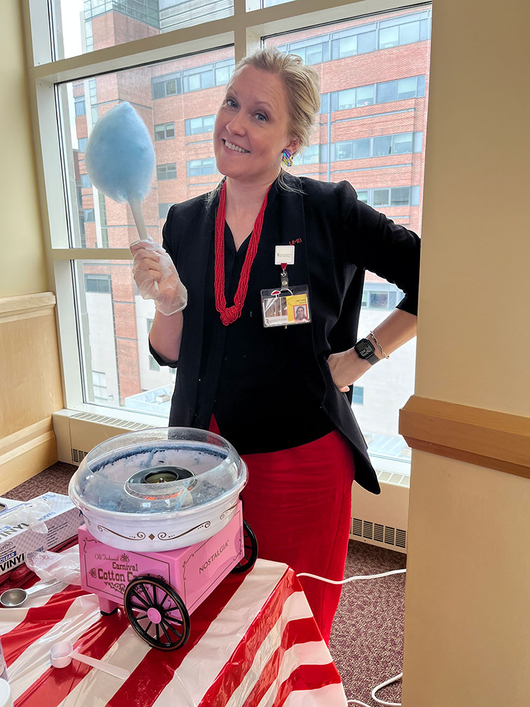 Emily Hurst holding blue cotton candy next to a pink cotton candy machine, with a brick building visible through the window.