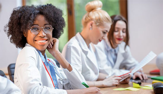 Three women in white medical coats collaborate at a table, with a smiling woman wearing glasses and a stethoscope in the foreground.