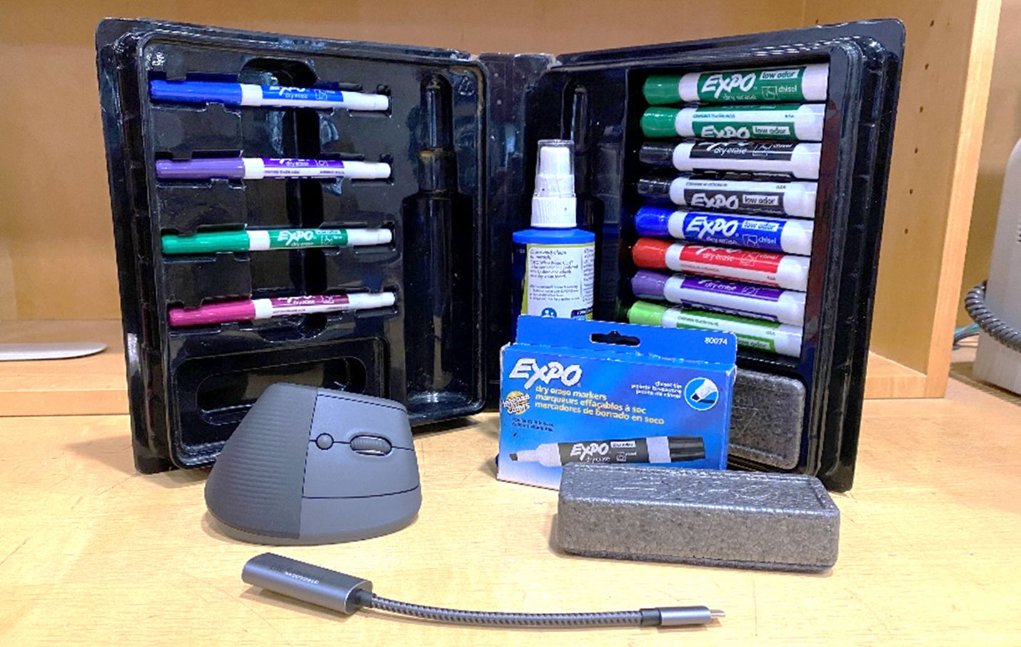 A collection of dry erase markers in an open case, with an ergonomic mouse, an Expo marker box, eraser block, and a USB gooseneck light on a wooden surface.