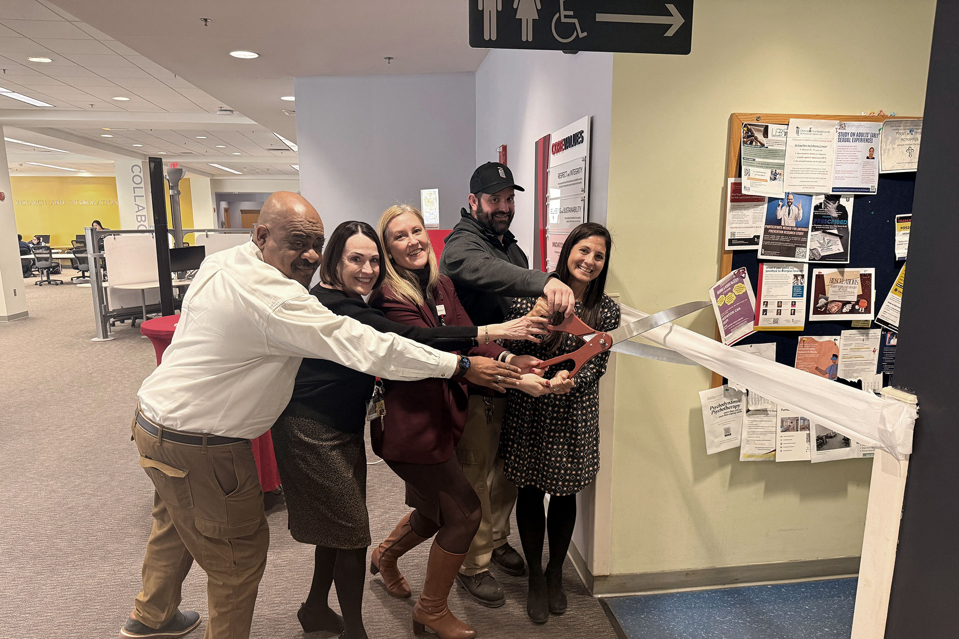 Five people cutting a white ribbon with large ceremonial scissors in a building corridor.