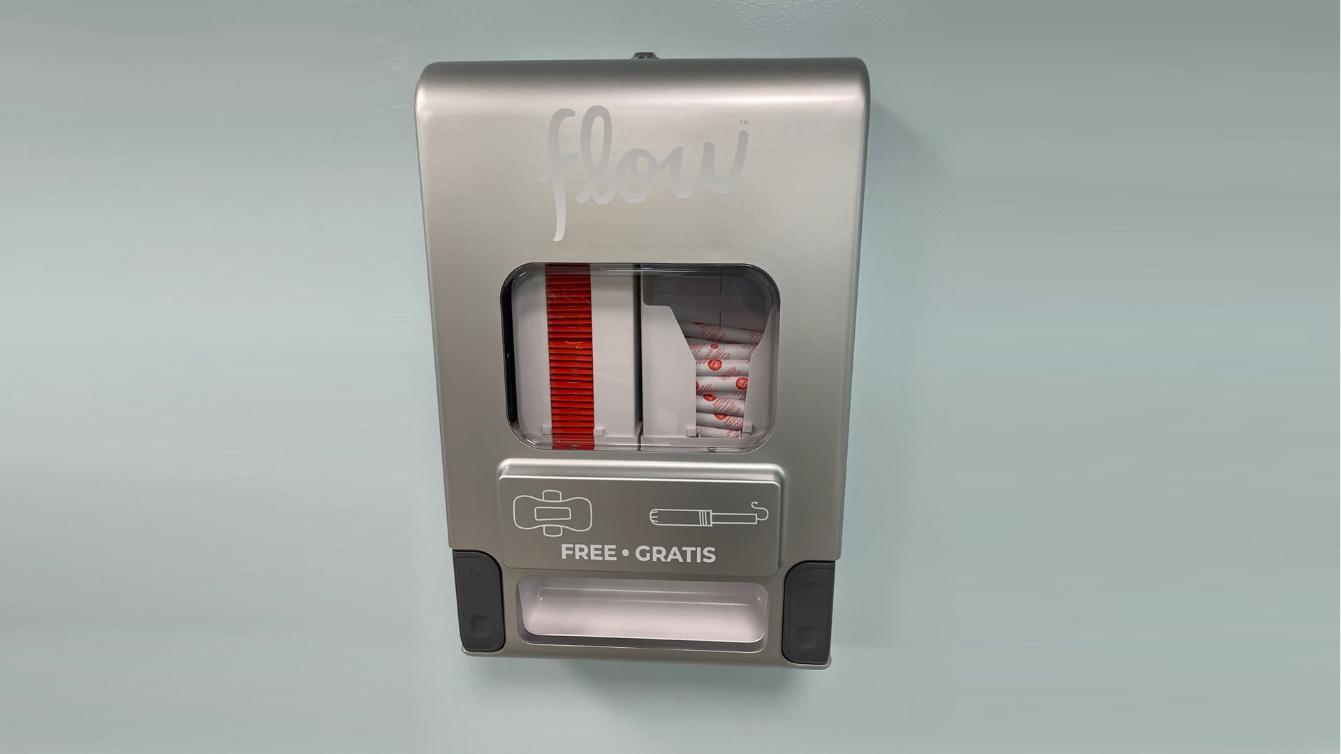 Silver feminine hygiene dispenser with FREE - GRATIS label on a light blue wall.