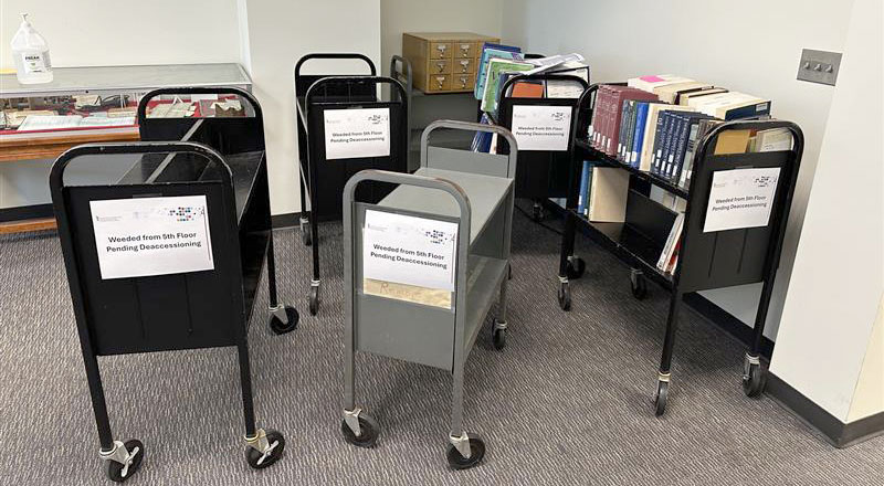 Several book carts with signs reading Weeded from 5th Floor Pending Deaccessioning are arranged in a library setting.