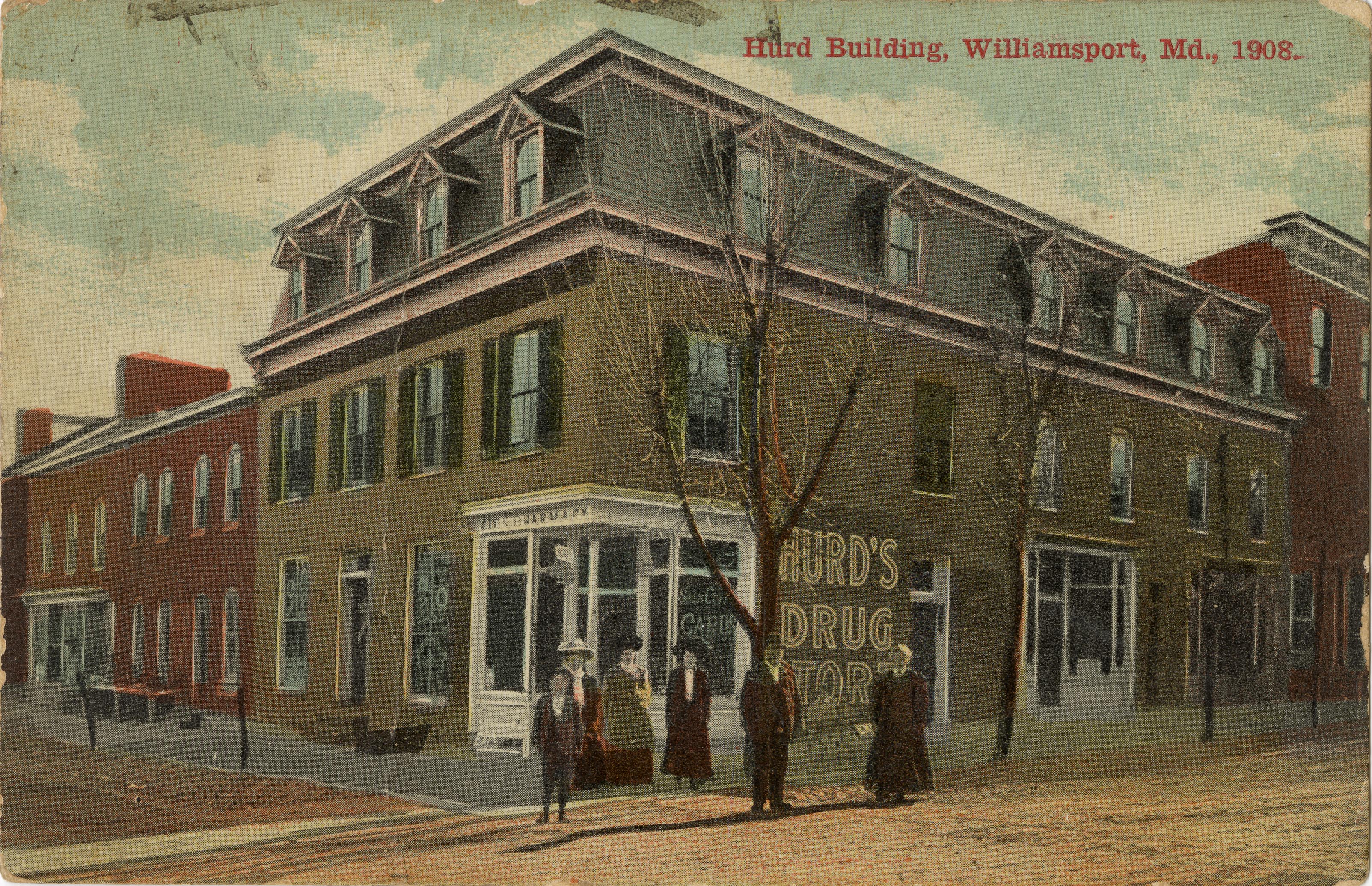 Postcard of a building on the corner of two streets, 6 people stand in front of the building. Building has Hurd's Drug Store on the side of it.