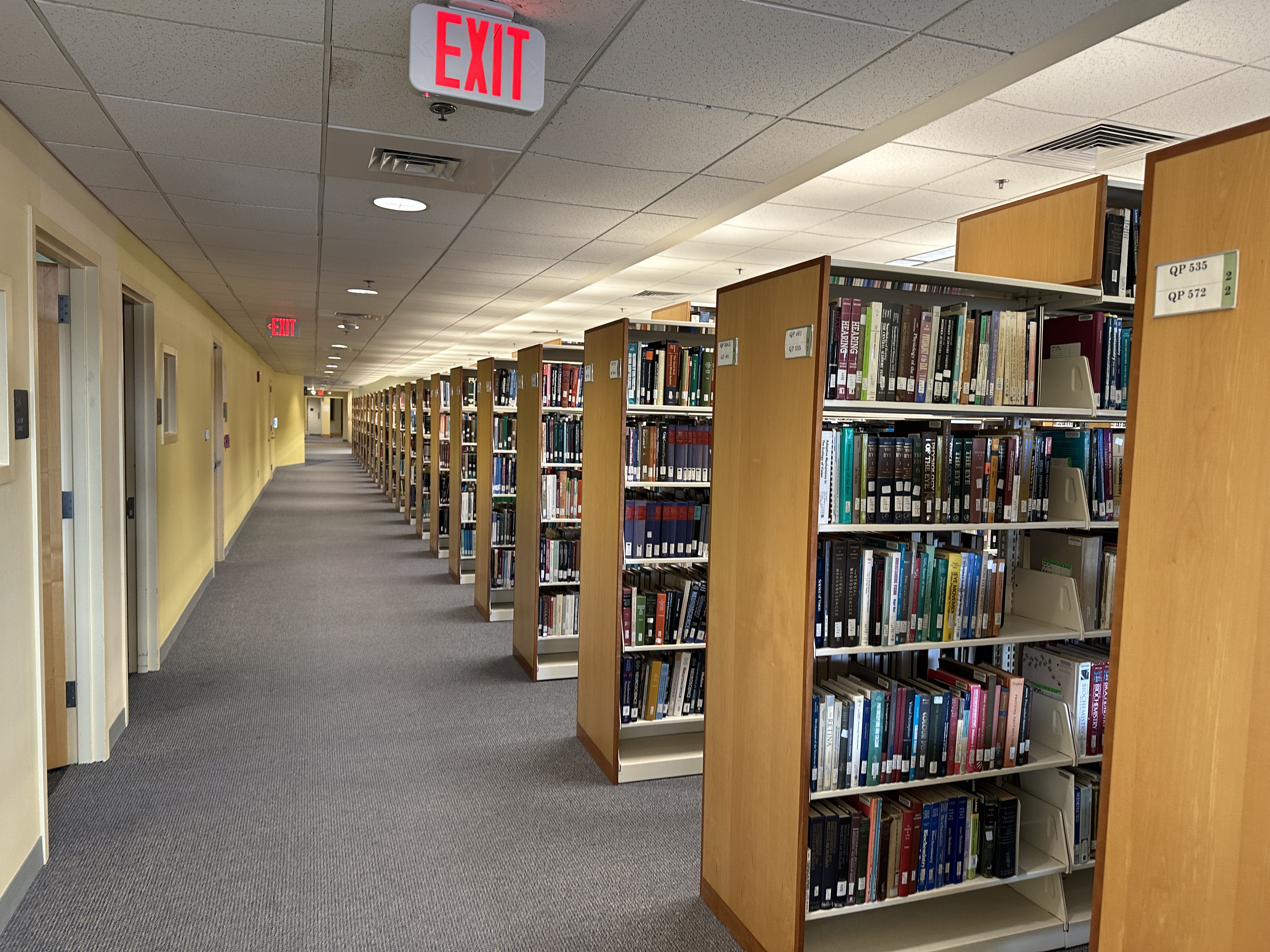 Photograph the fifth floor of the HSHSL. Photograph shows a long library aisle lined with tall wooden bookshelves filled with books, extending toward a distant exit sign. Ceiling lights illuminate carpeted floor and pale yellow walls.