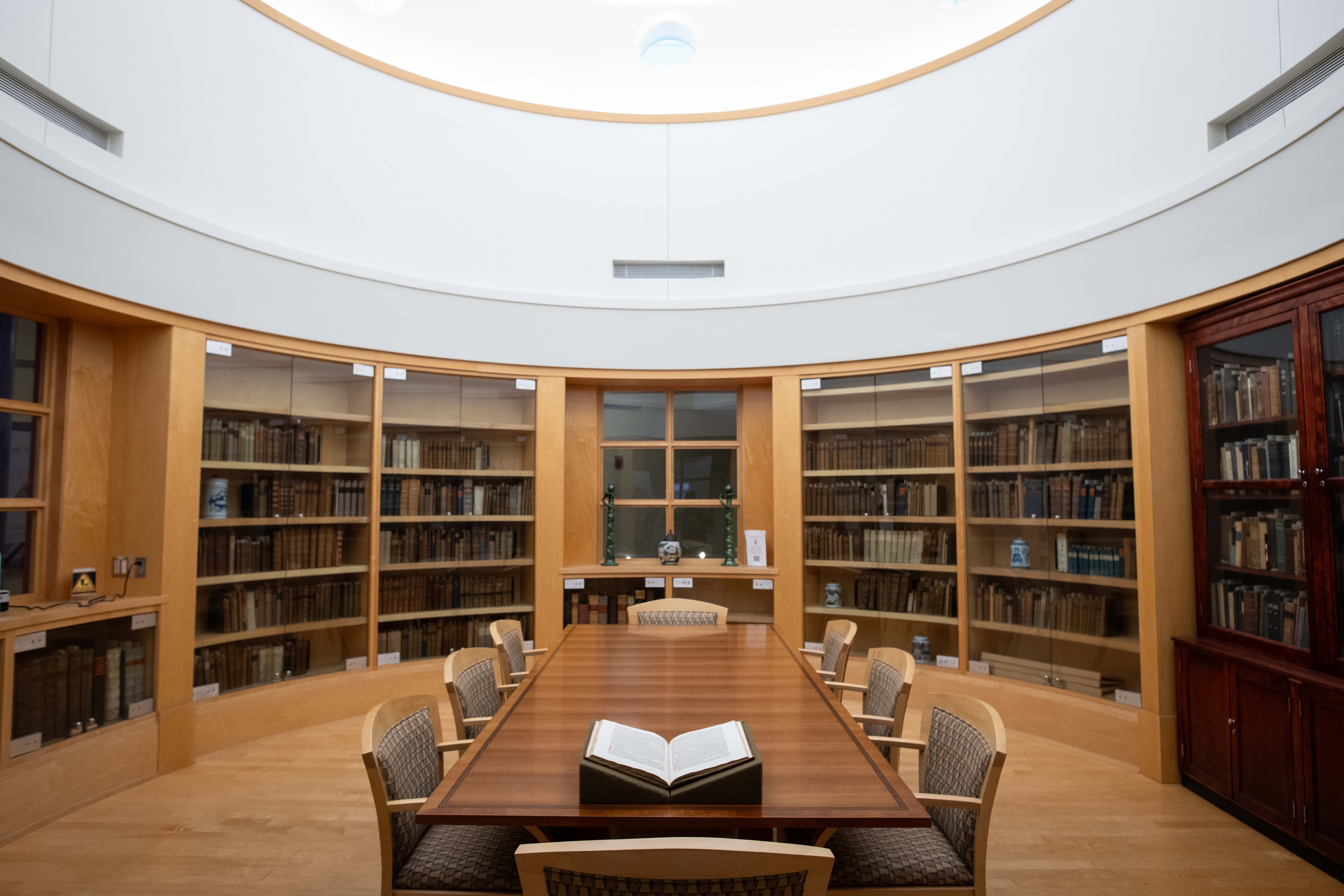 Photograph of a circular library reading room with curved wooden bookshelves filled with books. Central conference table, open book, chairs, glass-front cabinets, and bright domed ceiling create a scholarly study setting.