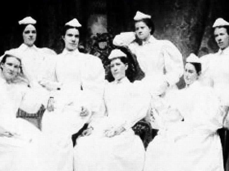 A historical black and white photo of eight female nurses in white uniforms and caps, arranged in two rows.